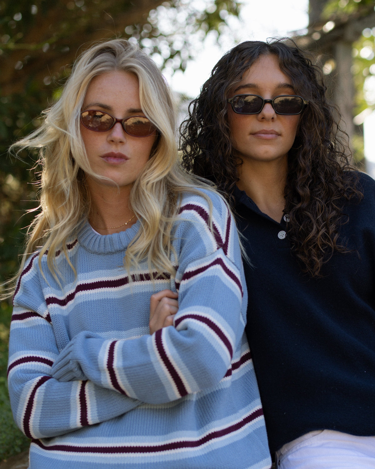 Two women sitting outdoors on a stone wall, wearing oversized knit sweaters. The woman on the left has blonde hair and wears a light blue sweater with dark red and white horizontal stripes, paired with denim shorts.
