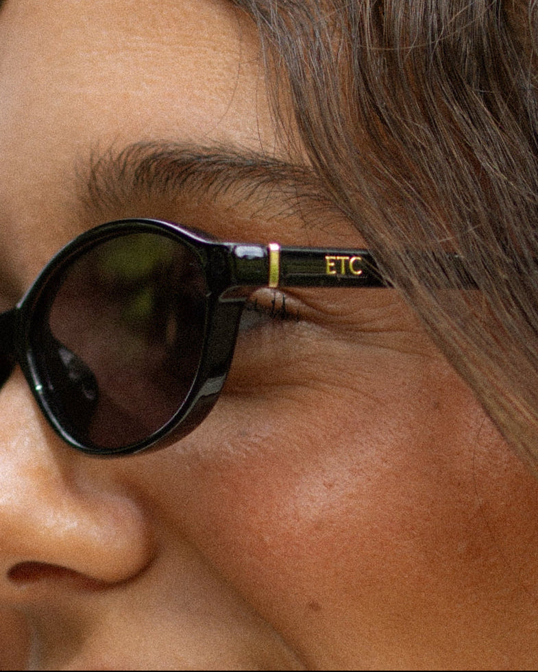 Close-up of a person wearing black oval sunglasses with gold lettering that reads 'ETC' on the temple arm.